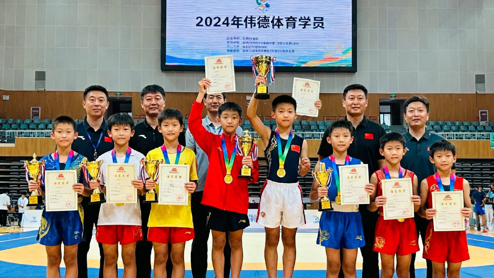 2024年伟德体育学员在多项省市比赛中获奖后的集体合影，十几位青少年选手穿着各色奖牌站在领奖台前，有的手持奖杯，有的举着证书，教练团队站在后排，背景是比赛场馆的巨大电子屏幕。